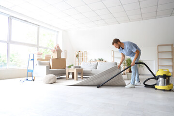 Male janitor cleaning floor under carpet with vacuum cleaner in room