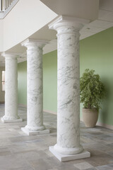 Three marble columns in an interior setting stand on a tiled floor beside a light green wall, with a potted plant nearby, reflecting a blend of modern and classical design