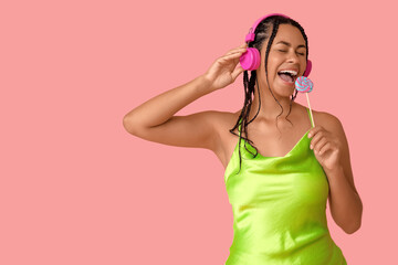 Young African-American woman with lollipop listening to music on pink background