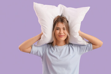 Young woman with pillow suffering from loud noise on lilac background