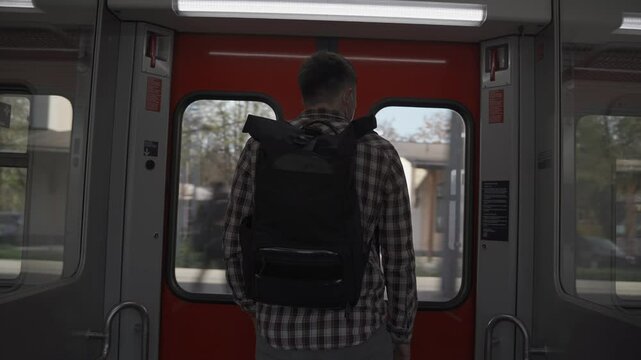 Man wearing backpack and shirt stands facing away near sliding doors of S-Bahn train in Munich, Germany. He waits for his stop to get off, depicting daily commuting in city public transport network. 