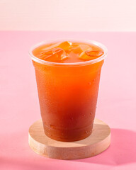 A cup of tea with ice cubes in it sits on a wooden coaster. The coaster is on a pink background