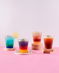A row of colorful drinks with a lemon slice in the middle. The drinks are served in plastic cups and are arranged on a pink background