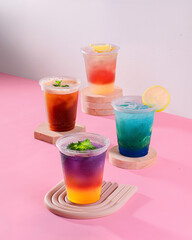 Four colorful drinks in plastic cups on a pink background. The drinks are in different colors and are arranged in a row