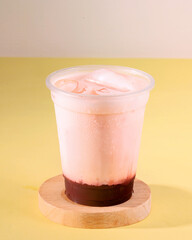 A cup of ice cream with a straw in it. The cup is on a wooden coaster. The ice cream is pink and...