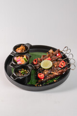 A plate of meat with a side of vegetables and a small bowl of sauce. The plate is black and the vegetables are green