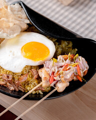 A bowl of food with a fried egg on top and a skewer of meat. The bowl is black and the food is colorful