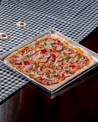 A pizza with pepperoni and cheese sits on a table. The pizza is cut into squares and is placed on a metal tray. The tray is on a checkered tablecloth