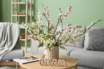 Vase of blooming branches with magazine and word DREAM on coffee table near sofa in living room, closeup