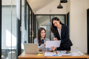Two women are sitting at a desk with a laptop and a stack of papers