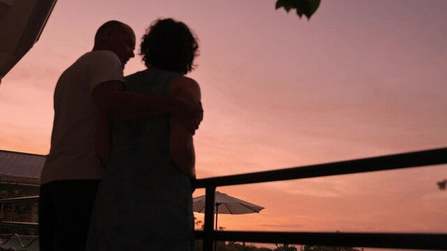 Silhouetted elderly couple standing close together on balcony during sunset, with soft pastel sky in background and gentle embrace capturing love, companionship, and peaceful tropical evening moment
