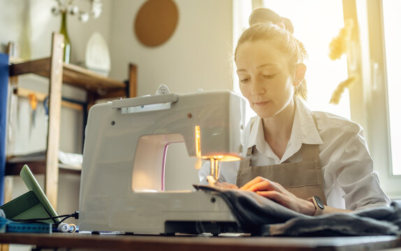 A female tailor is working on a sewing machine stitching fabric in sunny room. Home sewing production
