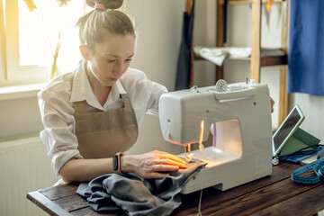 A female tailor is working on a sewing machine stitching fabric in sunny room. Home sewing production