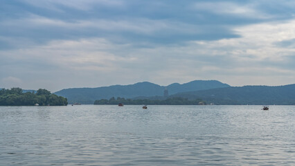 Tourist boats sail on the calm and beautiful lake. Ripples on the water. Green vegetation on the shore. The pagoda on the hill. Mountains against sky and clouds. China. Hangzhou. West Lake Xi Hu