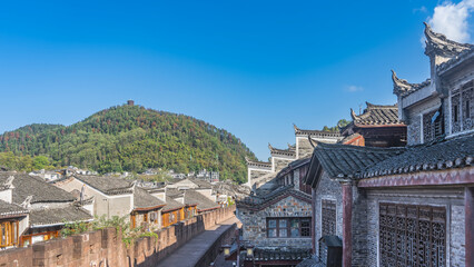 Naklejka premium The old fortress wall in the Fenghuang Ancient Town. Brickwork, walking path. There are many traditional Chinese buildings with curved tiled roofs. A green hill against a blue sky. China.