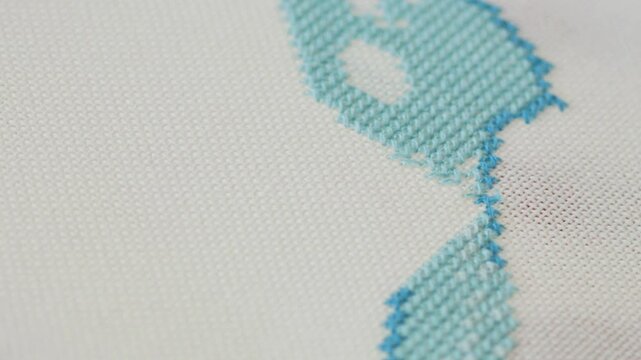 A close-up of a person hand skillfully stitching a light blue thread onto a textured white fabric, showcasing precision in needlework and craftsmanship. The process of cross-stitch embroidery