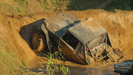 A muddy off-road vehicle designed for extreme off-roading partially peeks out of a deep puddle. The rear part of the vehicle is in water. There is a steep dirt slope in front of it.