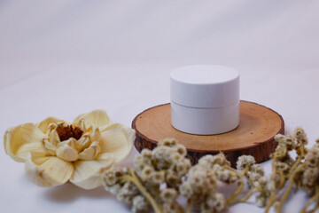 A minimalist and serene flat lay composition featuring a white, round cosmetic jar or container resting on a natural, circular wooden.