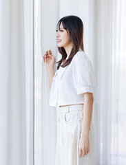 Smiling asian young beautiful pretty happy woman looking out opening curtains in morning and enjoying sunlight. Attractive female wearing white shirt white pants standing near bedroom window at home.