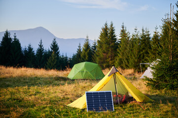 Solar panel in grassy field with tents in background highlights eco-friendly camping setup,...