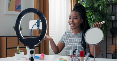 Beauty influencer uses ring light and phone tripod, filming tutorial on matching foundation to skin tone, desk full of makeup products, creating engaging content for followers.