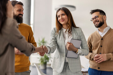 Female chief shaking hands with business partner while standing with her team of experts at office.