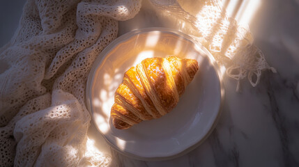 golden croissant sits on white plate, surrounded by soft, textured fabric, with sunlight casting gentle patterns. scene evokes warmth and comfort