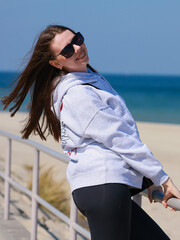 Young girl, with dark long hair, smiling, dark glasses, gray sweater, beach and sea behind