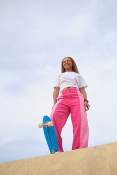 Young woman standing with skateboard at skatepark looking up smiling
