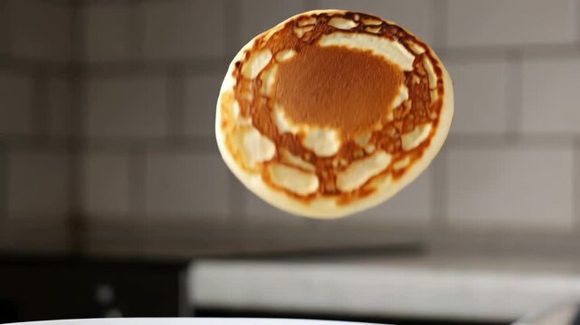 Slow-motion shot at eye-level captures a single, golden-brown pancake being deftly flipped high in the air by an unseen spatula against a clean kitchen background