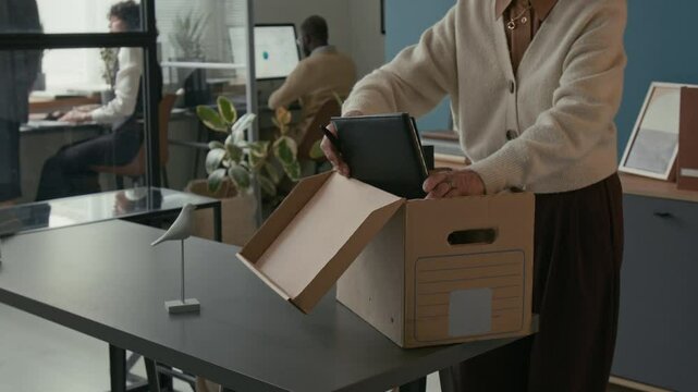 Cropped shot of Caucasian senior woman placing documents and folders into cardboard box as packing up her workspace during office retirement while colleagues working in background