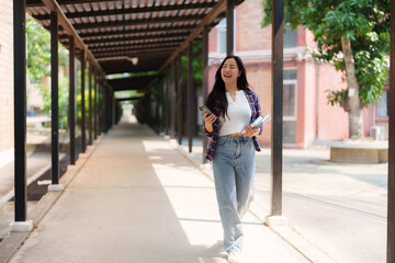 Happy asian female student walking through a university corridor, using her smartphone while carrying books, radiating positivity and embracing the vibrant campus atmosphere