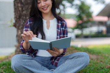 Fototapeta premium Young asian woman enjoys reading a book while sitting on grass in a park, turning a page with a smile, showcasing the joy of learning and leisure