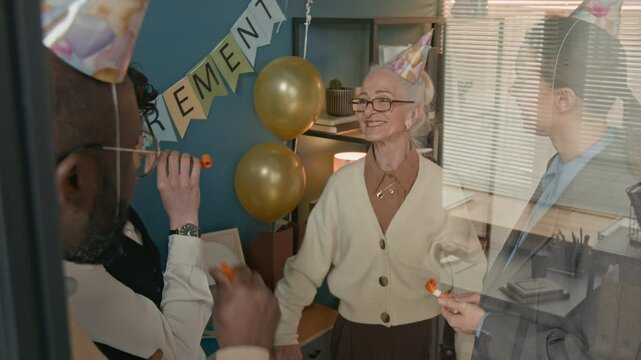 Team members joyfully blowing into party horns during cheerful retirement party of their senior colleague, surrounding smiling retiree with celebration, balloons, and laughter