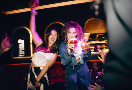 Two women celebrating with drinks in hand at a vibrant nightclub - Powered by Adobe