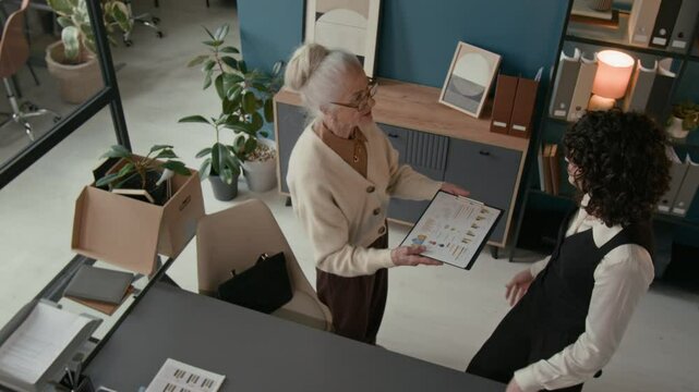 Overhead view of retiring woman going over important charts or information with her younger colleague on her last day in modern office