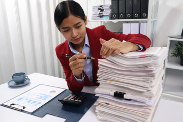 A young professional woman, examining a tall stack of documents with a focused expression.background are filled with more documents and black binders, suggesting a busy office environment