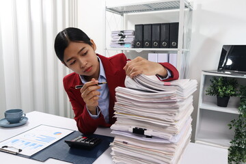 A young professional woman, examining a tall stack of documents with a focused expression.background are filled with more documents and black binders, suggesting a busy office environment