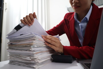 A young professional woman, examining a tall stack of documents with a focused expression.background are filled with more documents and black binders, suggesting a busy office environment
