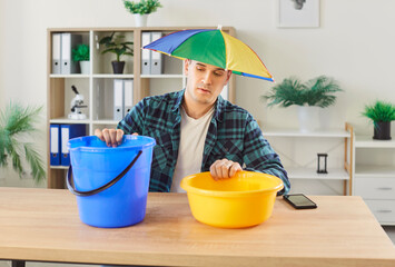 Troubled man at home in rainbow umbrella hat, hole or crack in ceiling through liquid flowing out...