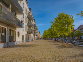 Charming view of Overgaden Neden Vandet along Christianshavn Kanal, Copenhagen. Cobblestone street, modern apartments, and lush trees under a clear blue sky. Ideal for urban and scenic themes.