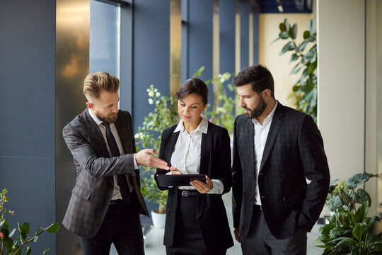 Business professionals in formal suits standing in office hallway, discussing collaborative project on tablet. Confident woman holding device while male colleagues analyzing data and sharing ideas. - Powered by Adobe