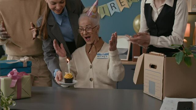 Medium shot of senior Caucasian woman wearing Retired badge smiling as she receiving cupcake with lit candle and wrapped gifts, surrounded by applauding coworkers at her office retirement party