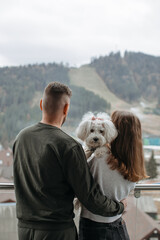 Couple hugging and holding their dog while enjoying mountain view from hotel balcony