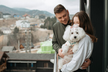Couple hugging and holding small white dog on balcony overlooking mountain village