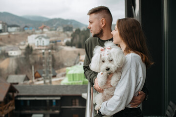 Couple hugging and holding small dog while enjoying mountain view from hotel balcony