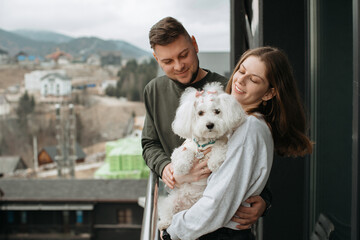 Happy couple embracing and holding small dog on hotel balcony with mountain view