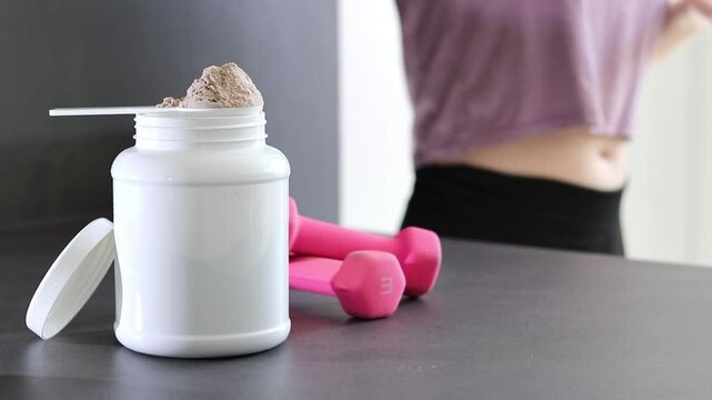 Close up of whey protein powder box and pink dumbbells on table, woman preparing a protein shake
