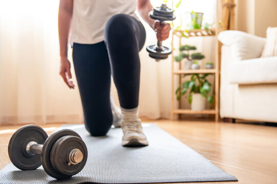 Mature woman exercising with dumbbells at home, kneeling on a yoga mat in her living room, demonstrating a commitment to fitness and a healthy lifestyle