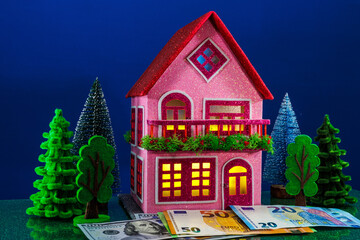 Euro and dollar banknotes lie in front of a pink house surrounded by trees and fir trees with yellow lights in the windows